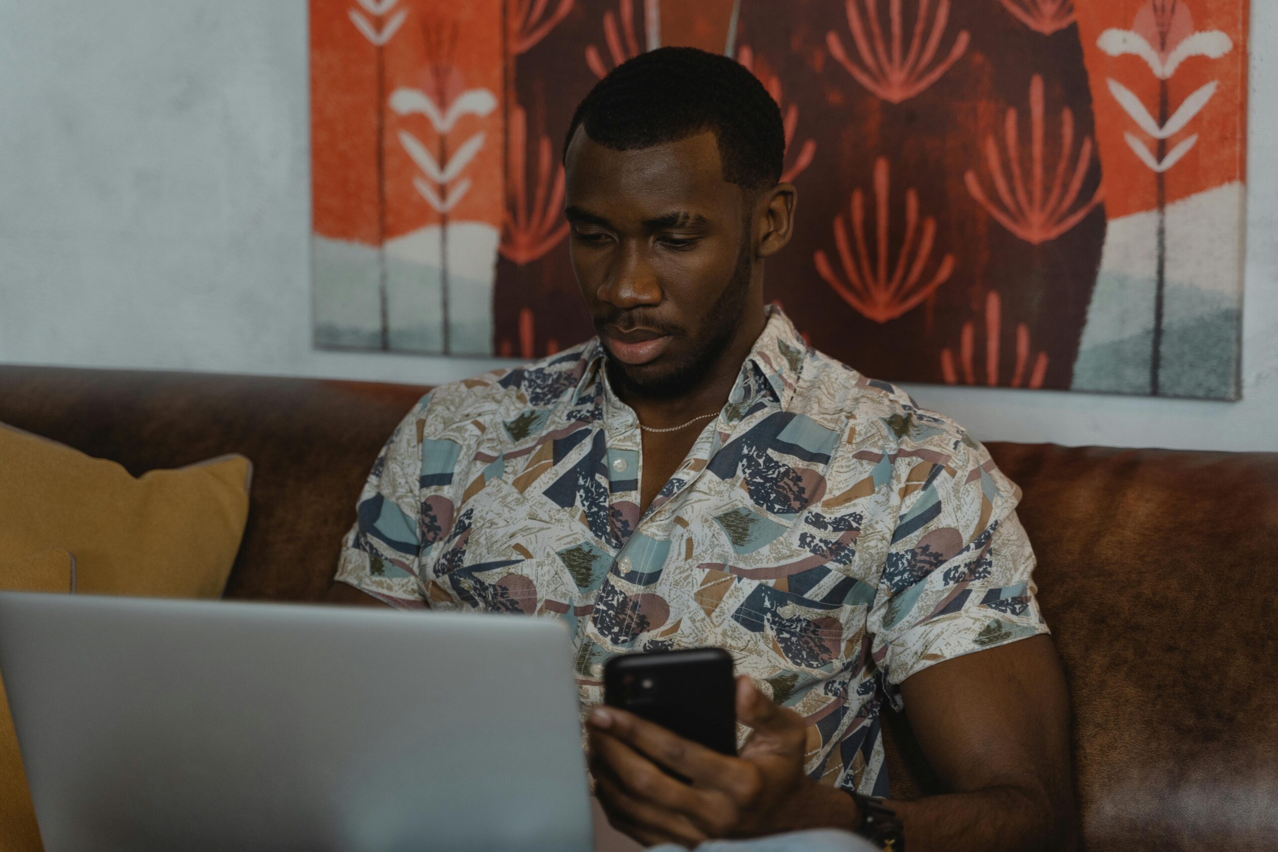 A man sitting on a couch working on a laptop while checking his smartphone.